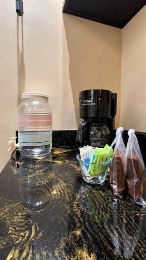 Uma bancada de cozinha com uma cafeteira, um bebedouro, um pote de vidro, uma tigela com sachês de café e um saco plástico contendo um objeto marrom.