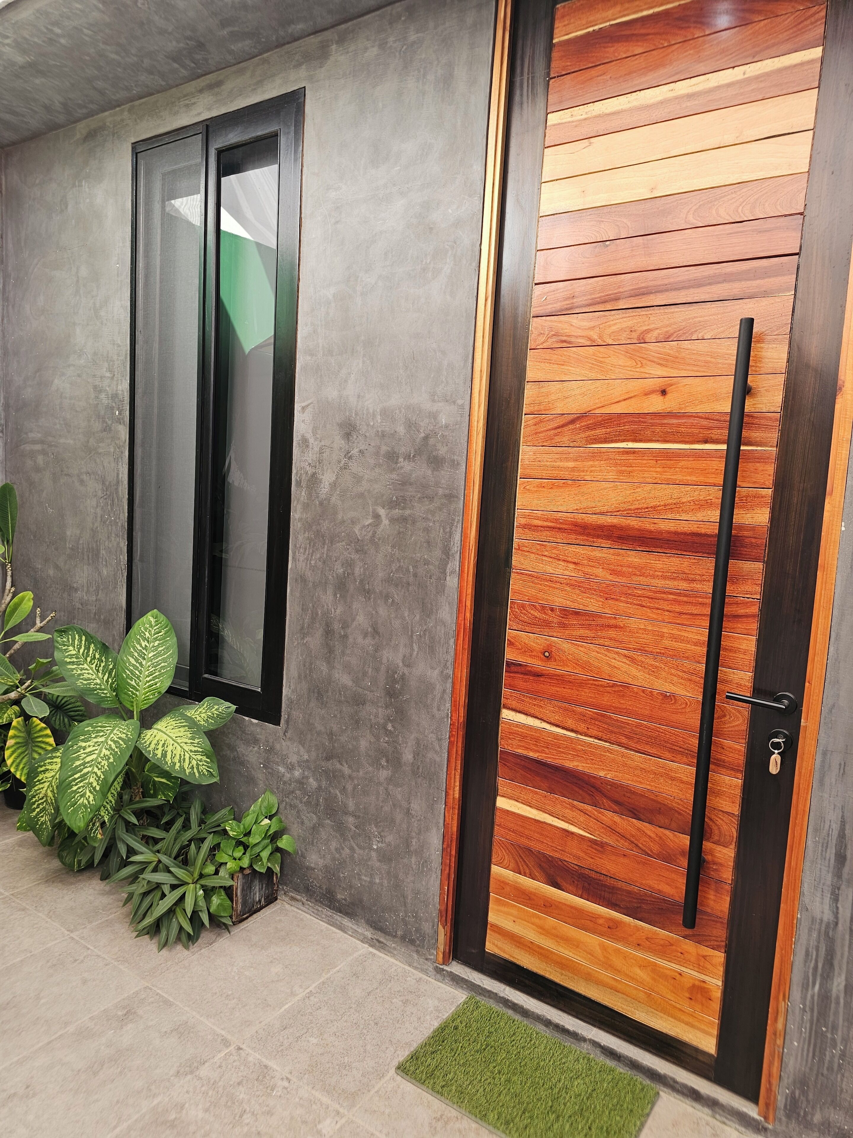 Une façade moderne avec un mur en béton, une porte en bois dotée d’une poignée en métal et un paillasson vert.