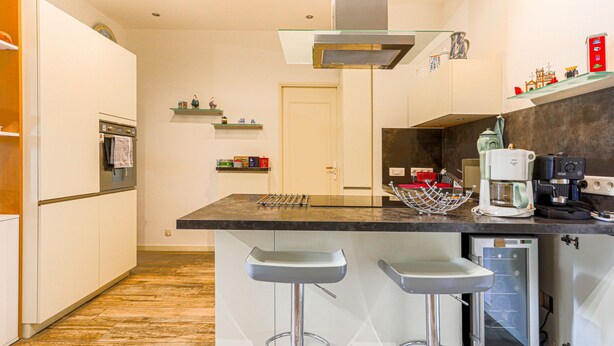 Een moderne keuken met een centraal eiland, barstoelen, een inbouwoven en een koffieapparaat.