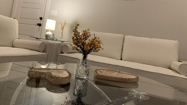 A modern living room with a glass coffee table, a white sofa, and a vase with yellow flowers. There are framed artworks on the wall and a lamp on a side table. - Cozy Southern Retreat, 4 Spacious Bedrooms, Massage Chair, 8 Mins to Ole Miss (Oxford)