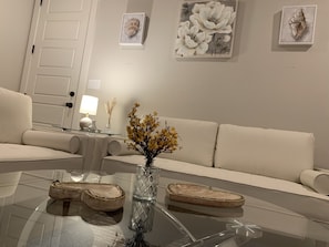 A modern living room with a glass coffee table, a white sofa, and a vase with yellow flowers. There are framed artworks on the wall and a lamp on a side table.