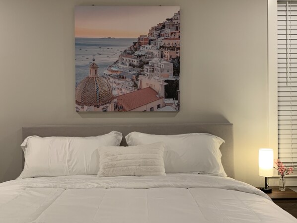 A bedroom with a large bed, a nightstand with a lamp, and a wall-mounted picture of a coastal town.