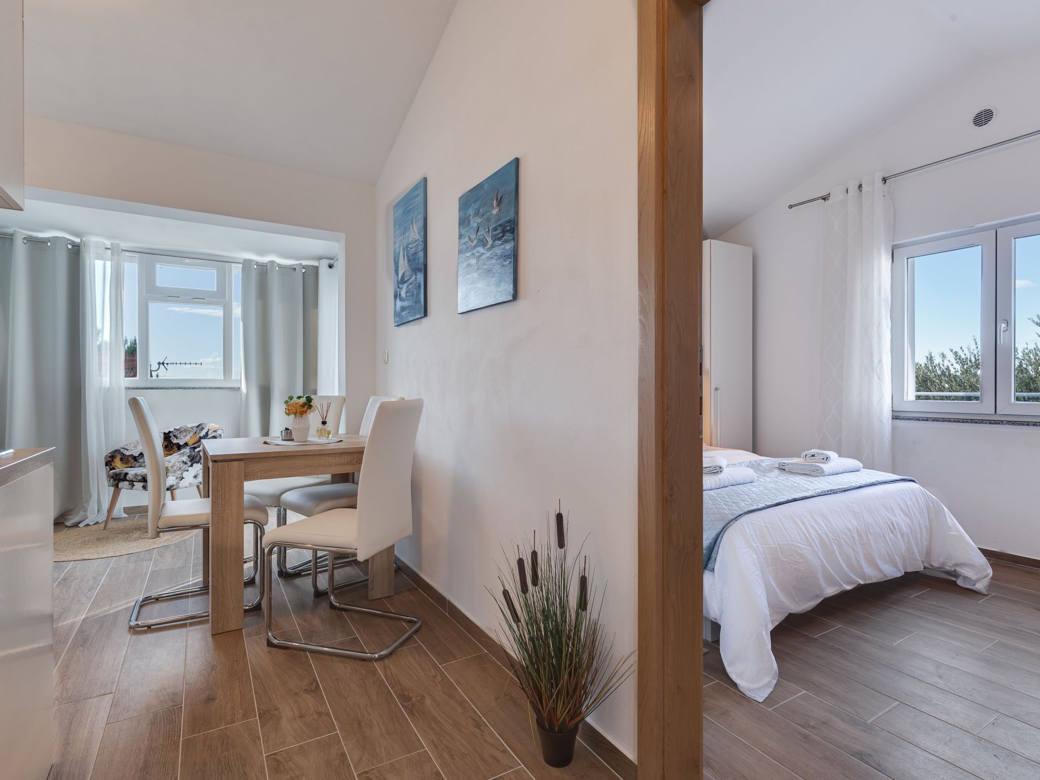 Un dormitorio moderno con cama, un escritorio pequeño y una silla. Hay una ventana con cortinas, una planta en maceta y un cuadro en la pared.