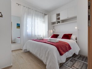 Ein modernes Schlafzimmer mit einem großen Bett, weißer Bettwäsche, roten Kissen, einem weißen Kopfteil und einem kleinen Regal darüber.