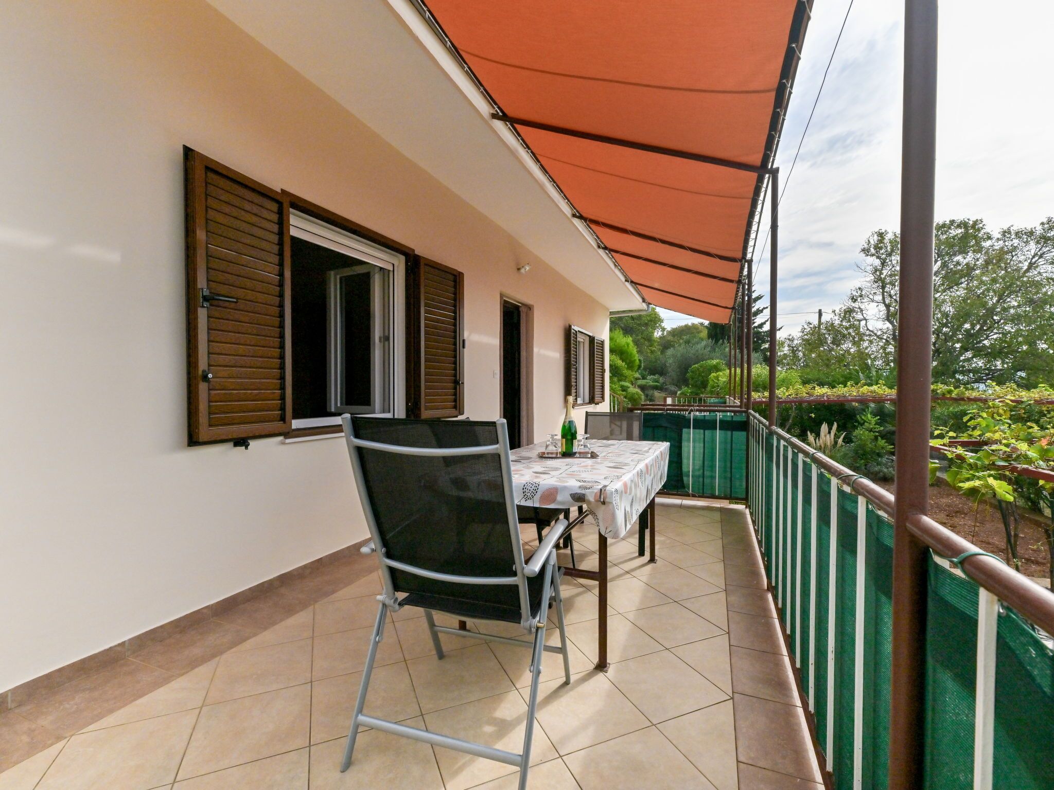 A covered balcony with a table and chairs, a window with shutters, and a view of greenery.