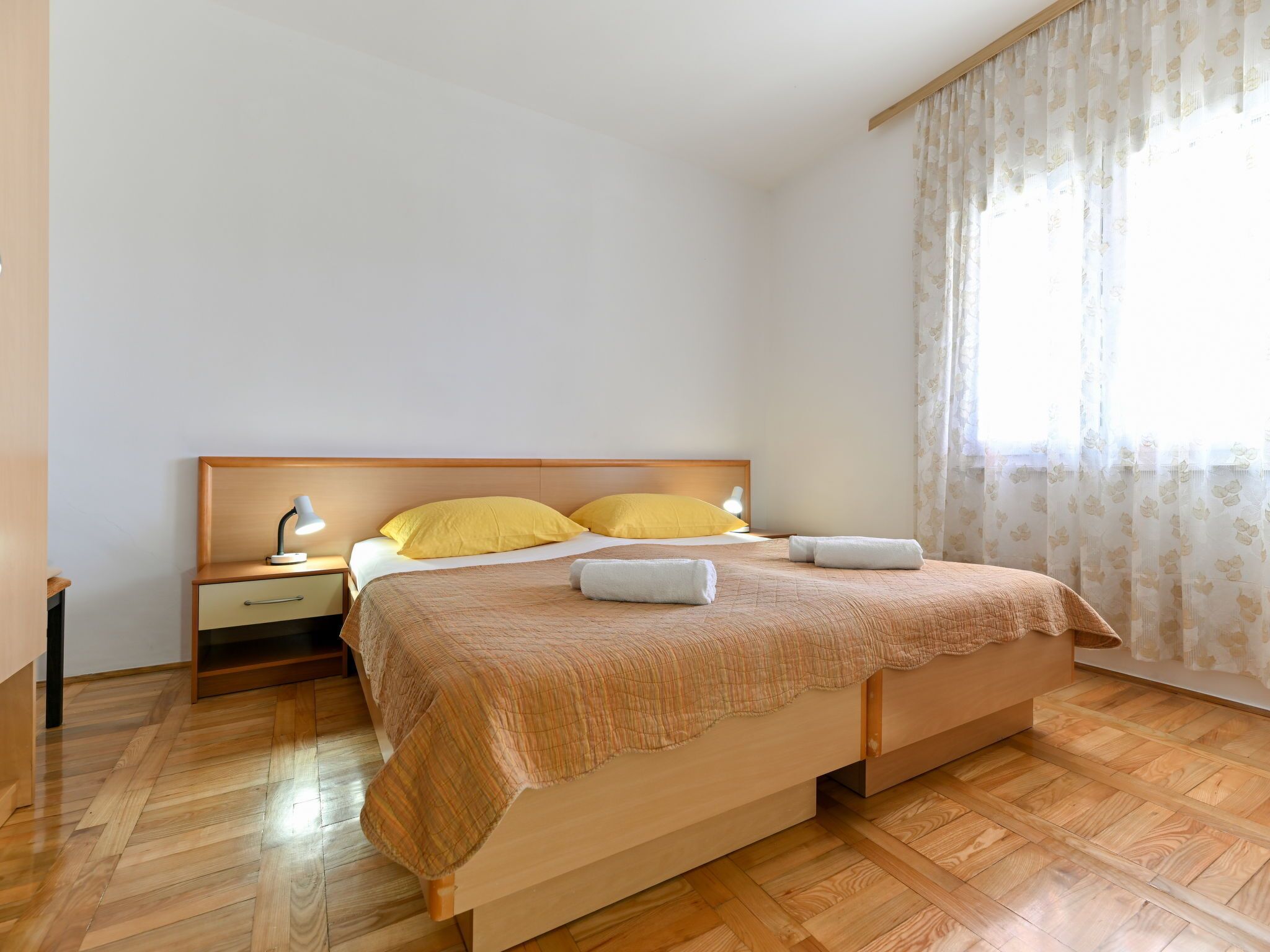 A bedroom with a wooden bed, two bedside tables with lamps, a window with curtains, and a wooden floor.