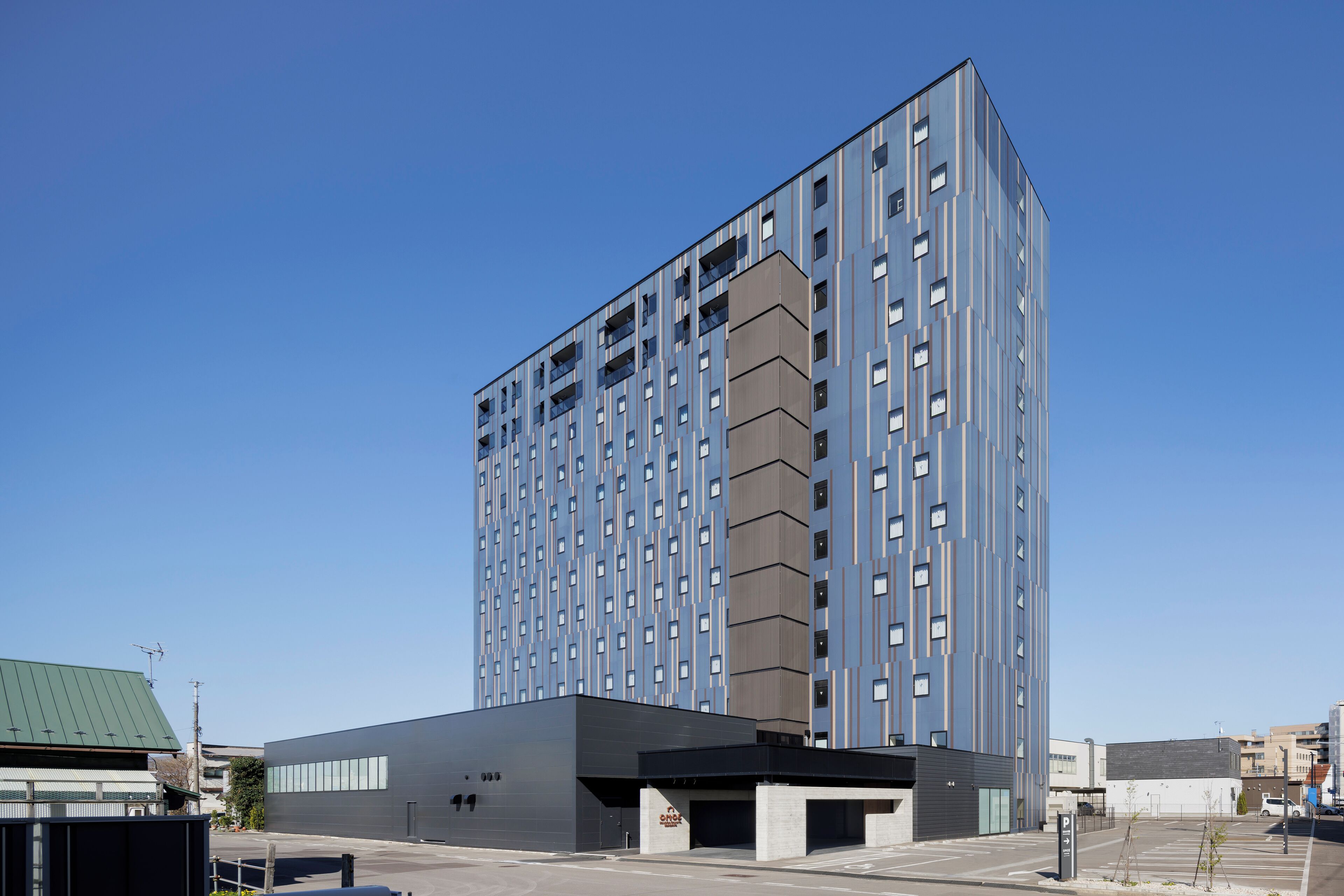 A modern high-rise building with a grid of windows and balconies, a flat roof, and a parking area in front.