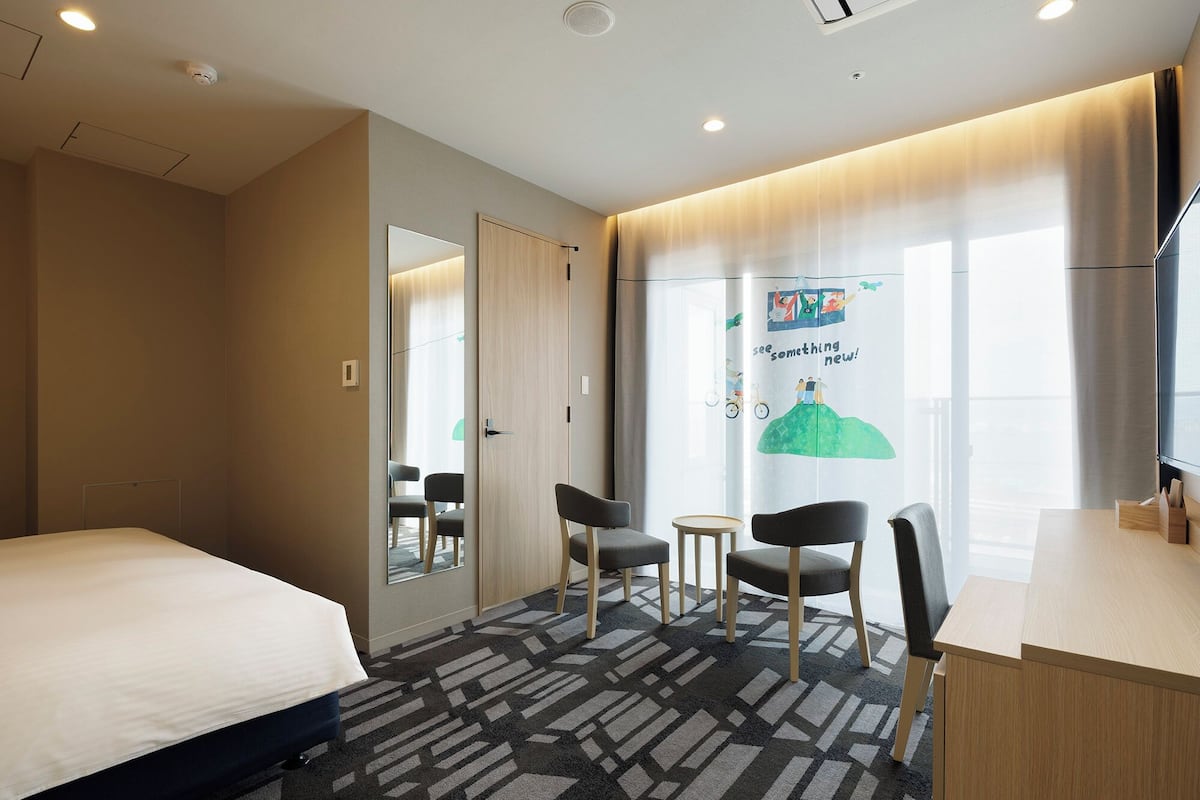 Une chambre d’hôtel avec un lit, deux chaises, une petite table, un bureau et une grande fenêtre avec des rideaux.