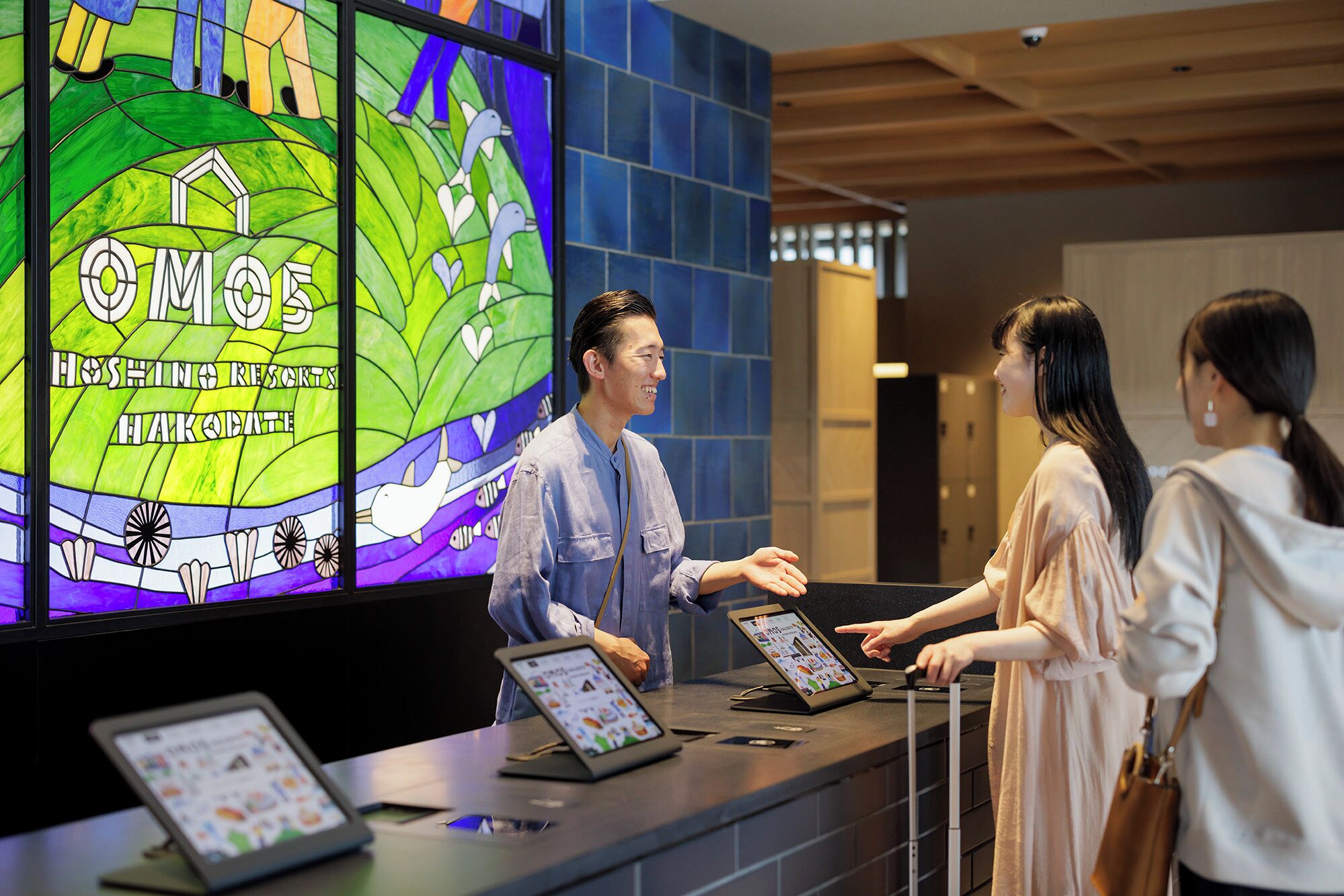 A reception area with a digital display featuring a colorful stained glass design and text, a counter with tablets, and a modern interior with blue tiles and wooden ceiling.