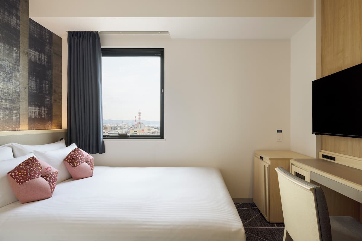 Une chambre d’hôtel équipée d’un lit, d’un bureau, d’une télévision et d’une fenêtre donnant sur la ville.