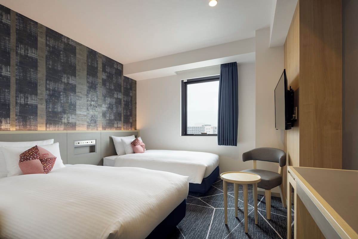 Une chambre d’hôtel avec deux lits, une petite table, une chaise, une fenêtre avec des rideaux et un téléviseur fixé au mur.