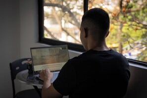 Una persona sentada a una mesa con una computadora portátil, mirando por una ventana con vistas a árboles y un edificio.