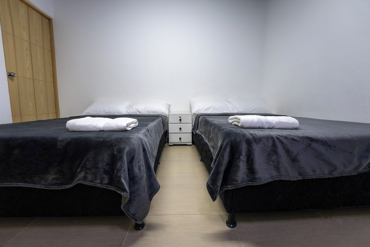 A room with two beds, each with a dark cover and white pillows, and a wooden wardrobe to the left.