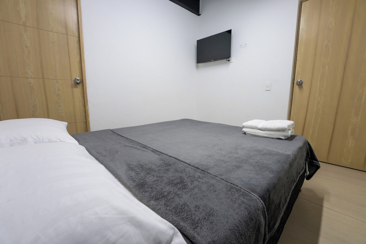 A bedroom with a bed, grey and white bedding, a flat-screen TV mounted on the wall, and wooden wardrobes.