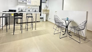 Une cuisine moderne avec des meubles blancs et un îlot central, un coin repas avec des chaises en fil de fer noir et une petite table ronde, et un plafond avec des poutres apparentes et un éclairage décoratif.