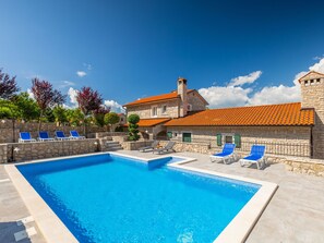 Une piscine avec des chaises longues, un bâtiment en pierre doté d’un toit en tuiles et un ciel d’un bleu limpide.
