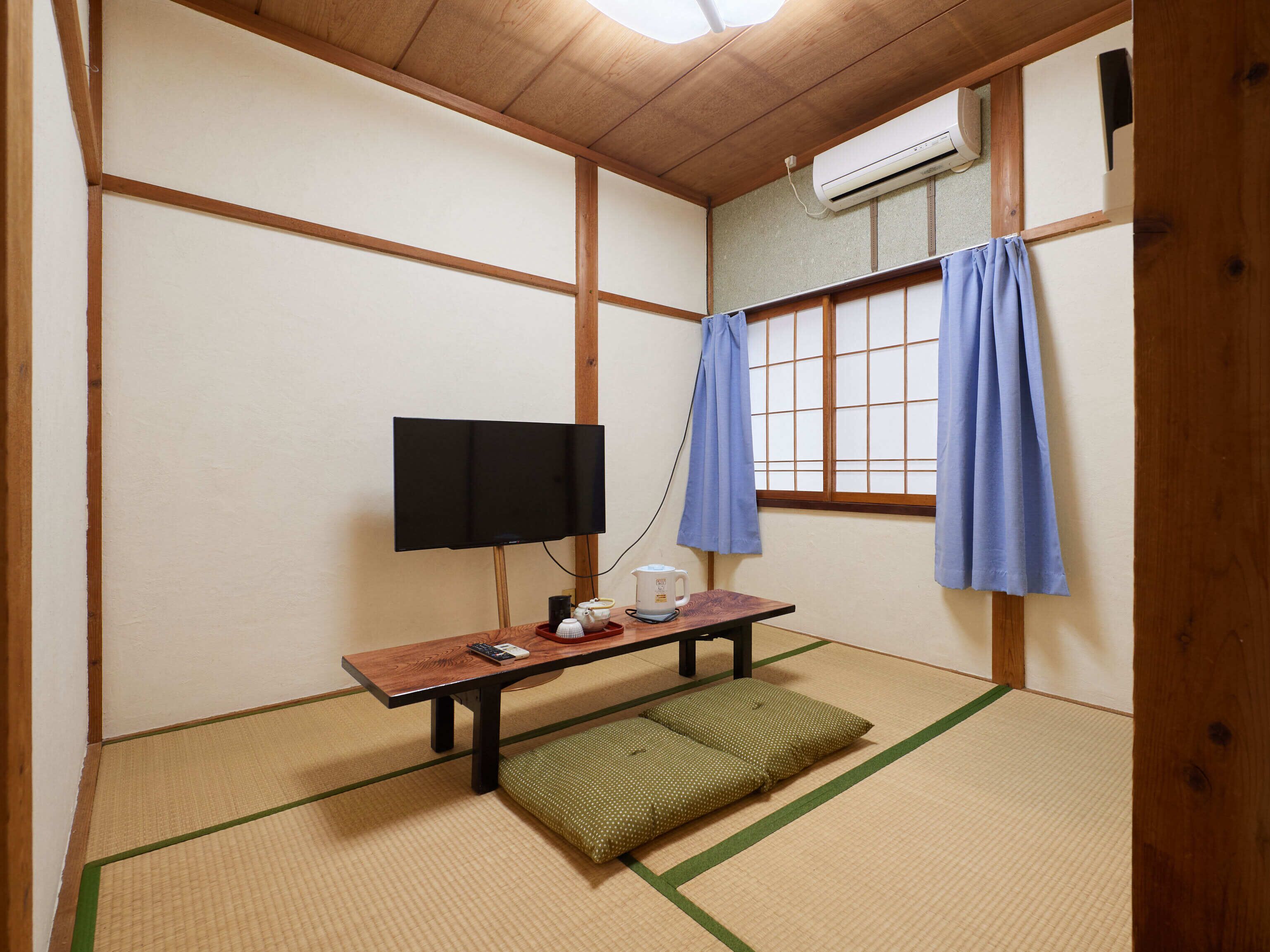 Una habitación tradicional japonesa con suelo de tatami, una mesa baja con televisor, un calentador y una ventana con cortinas azules.