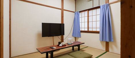 Una habitación tradicional japonesa con suelo de tatami, una mesa baja con televisor, un calentador y una ventana con cortinas azules.