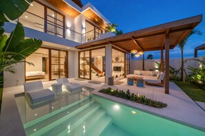 Una casa moderna de dos pisos con piscina cubierta de vidrio, mobiliario exterior y un patio cubierto.