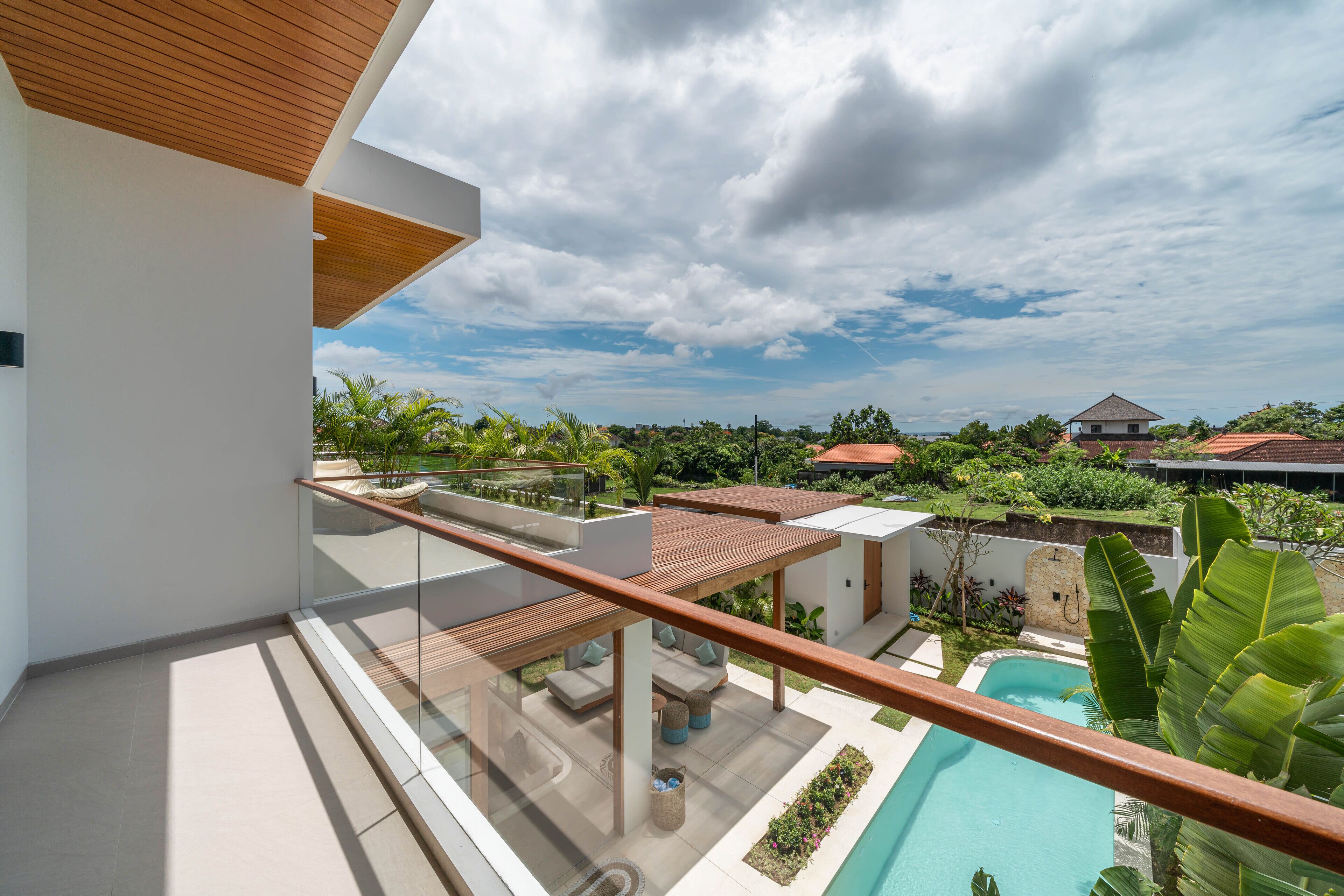A modern outdoor balcony with a wooden ceiling, a glass railing, and a view of a swimming pool and lush greenery.