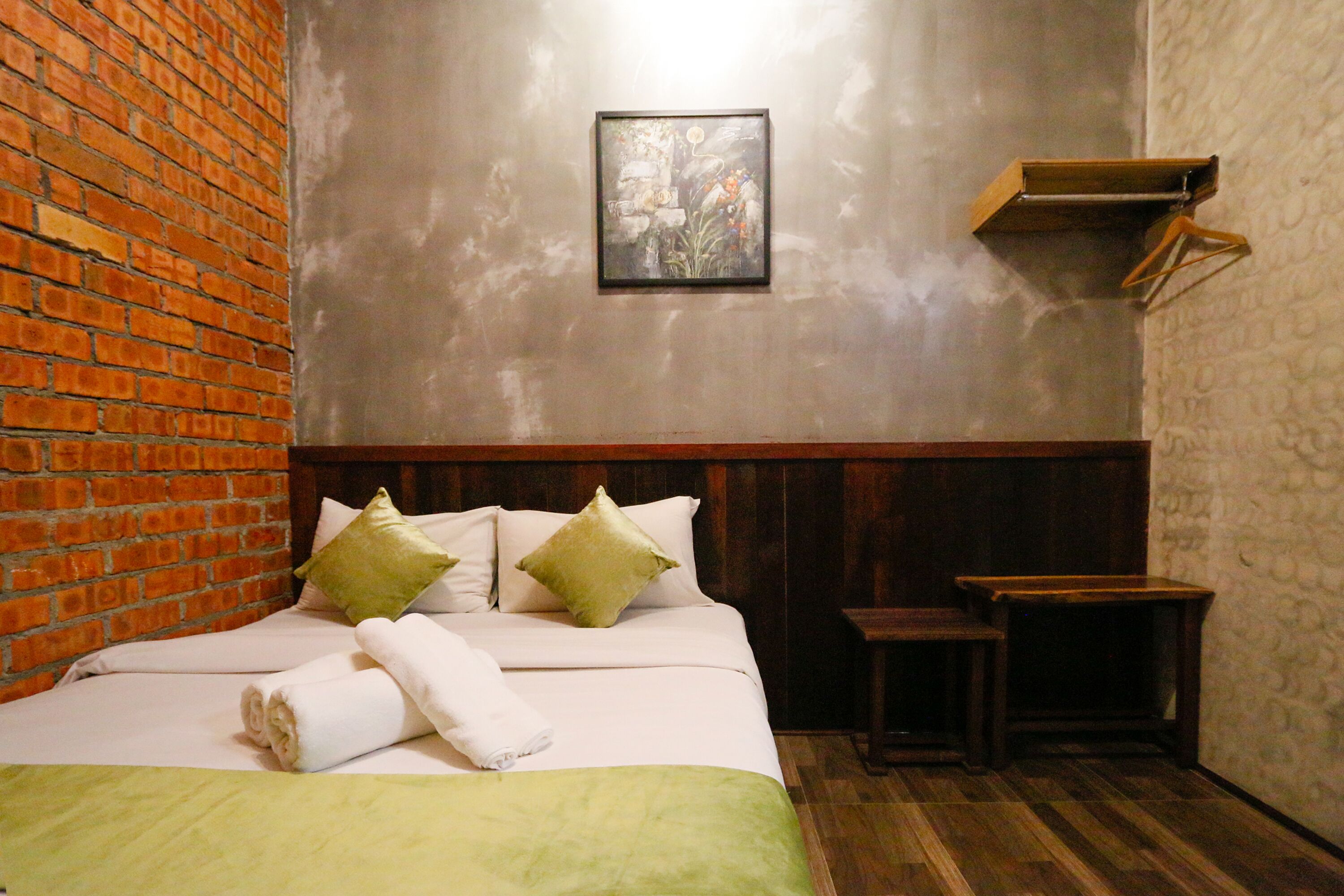 A hotel room with a brick wall, a bed with green pillows, a framed picture on the wall, and a small wooden table.