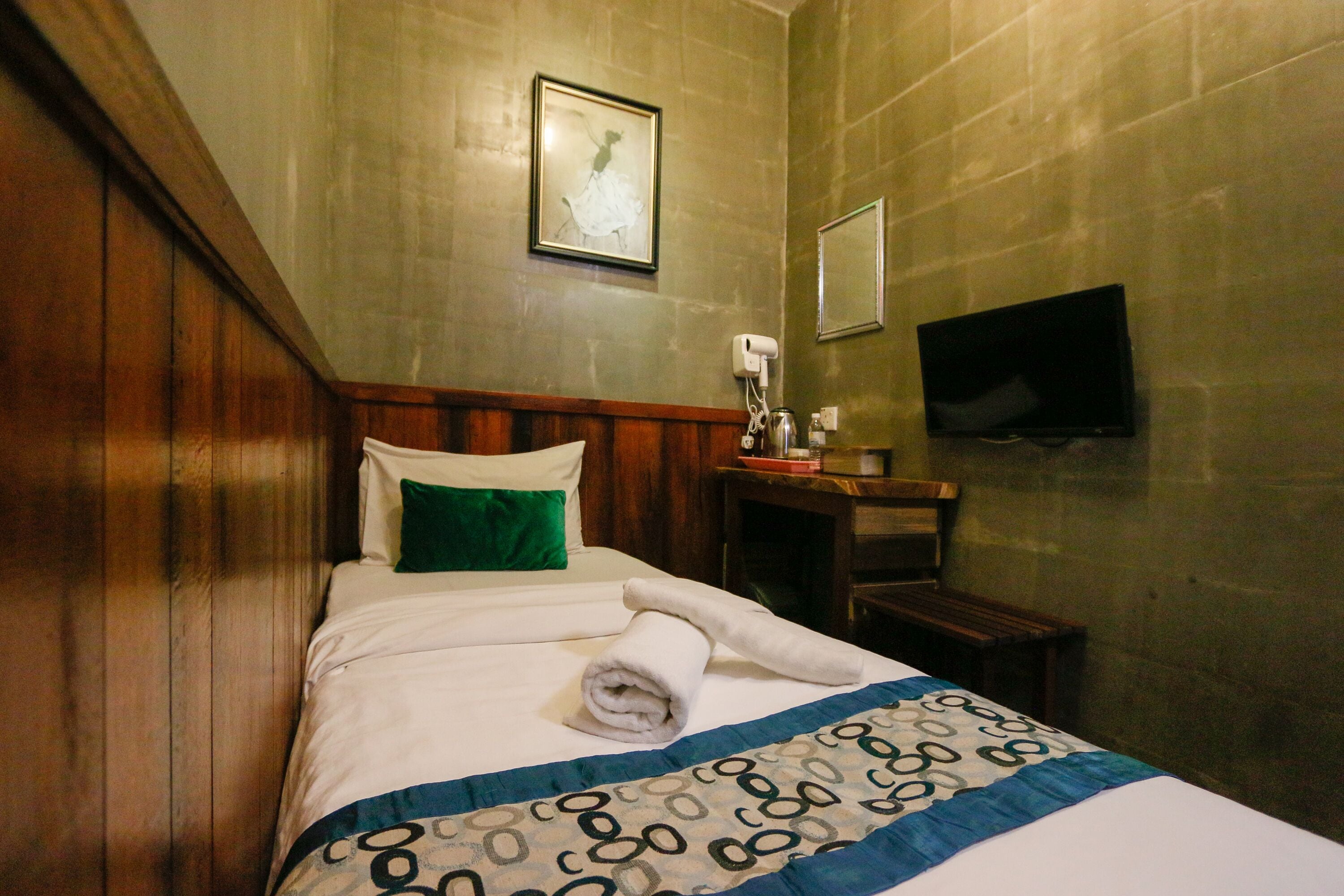 A hotel room with a wooden headboard, a bed with a patterned bedspread, a green pillow, a rolled towel, a bedside table with a lamp, a framed picture on the wall, and a flat-screen TV mounted on the wall.