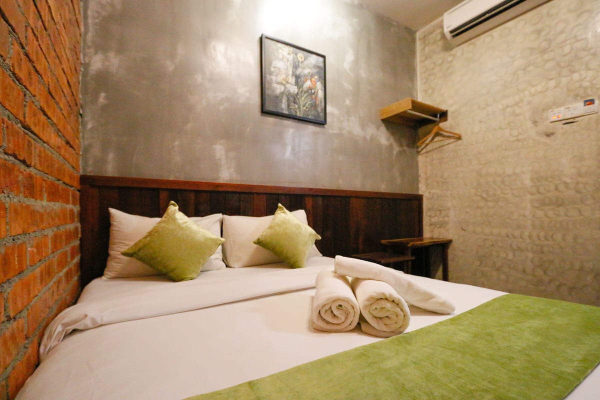 A hotel room with a brick wall, a bed with white bedding and green pillows, a framed picture on the wall, and a small wooden nightstand.