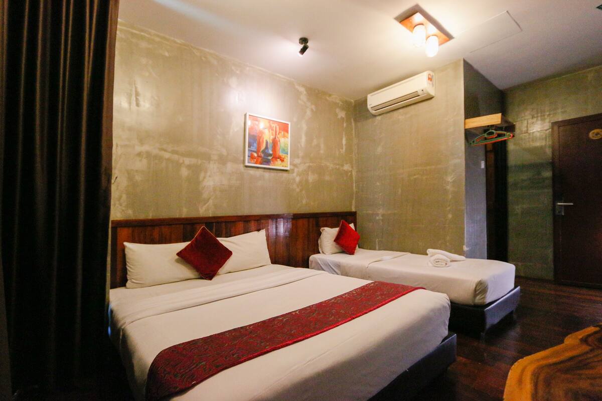 A hotel room with two beds, a red throw, a painting on the wall, and an air conditioning unit.