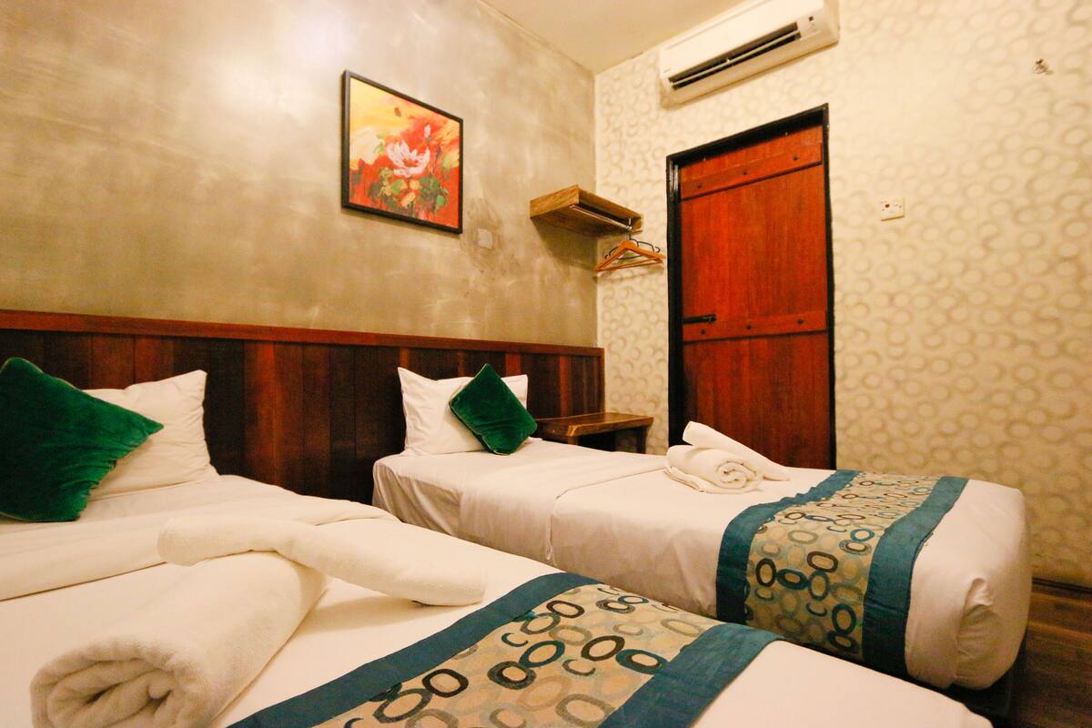 A hotel room with two beds, each with a pillow and a rolled towel. There is a painting on the wall and an air conditioning unit above the door.