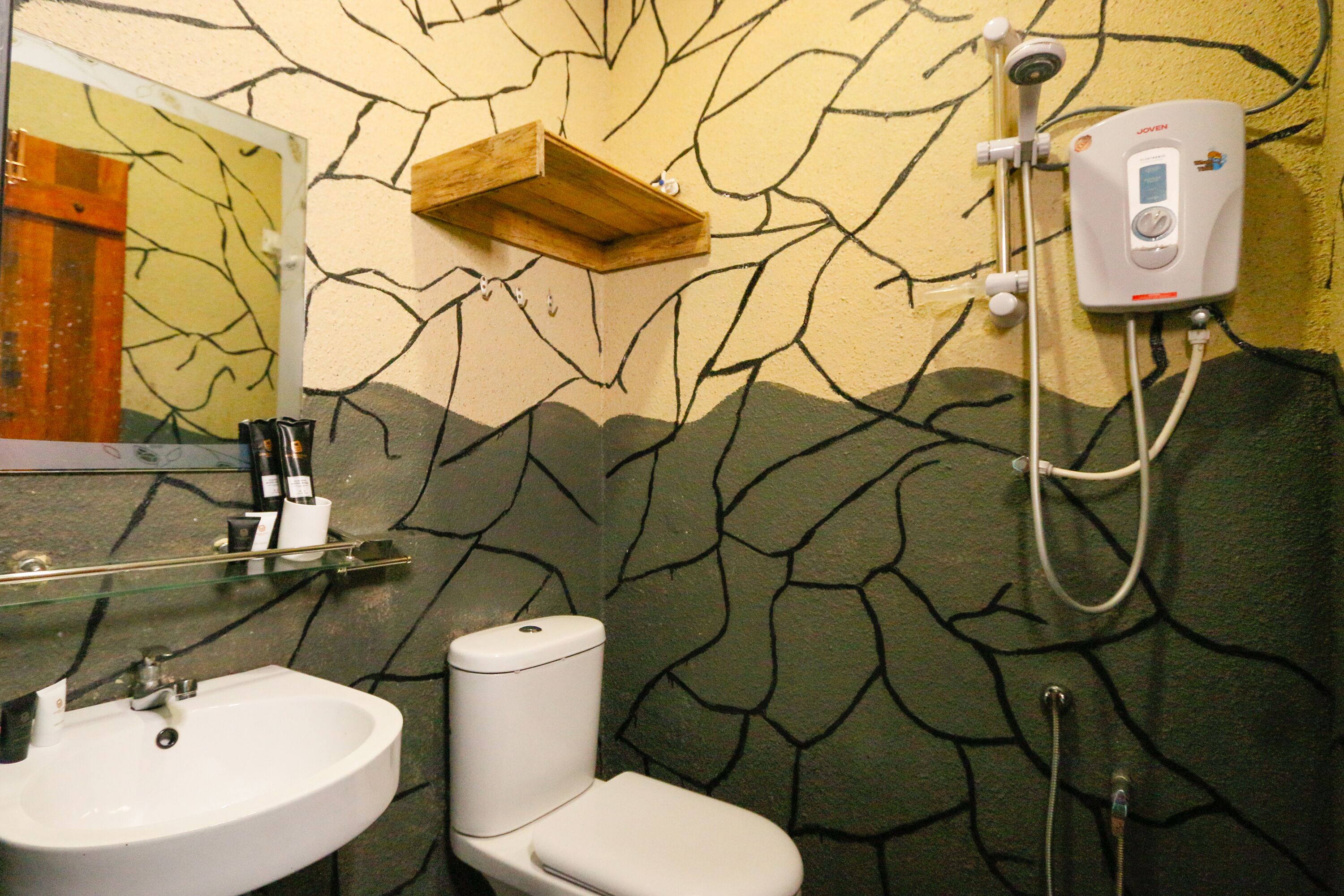 A bathroom with a white sink, toilet, and shower. There is a wooden shelf above the toilet and a mirror on the wall.
