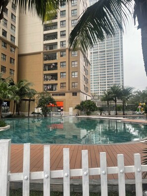 A swimming pool with a wooden deck, surrounded by a white fence, and adjacent to high-rise buildings.