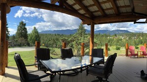 A covered outdoor seating area with a glass table and chairs, overlooking a mountainous landscape. - LAKE VIEW Mountain house w/ HOT TUB
 (Invermere)