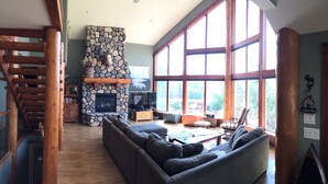 A spacious living room with a stone fireplace, large windows, and a wooden staircase. - LAKE VIEW Mountain house w/ HOT TUB
 (Invermere)