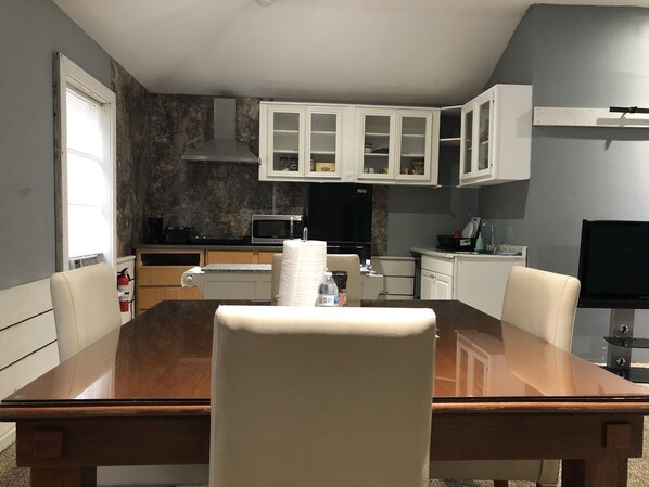 A dining area with a wooden table and chairs, a kitchen with white cabinets, a microwave, and a black TV.