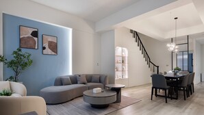 Una sala de estar moderna con un sofá modular, una mesa de centro redonda y un comedor con mesa y sillas. Se ve una escalera que sube a otro nivel.