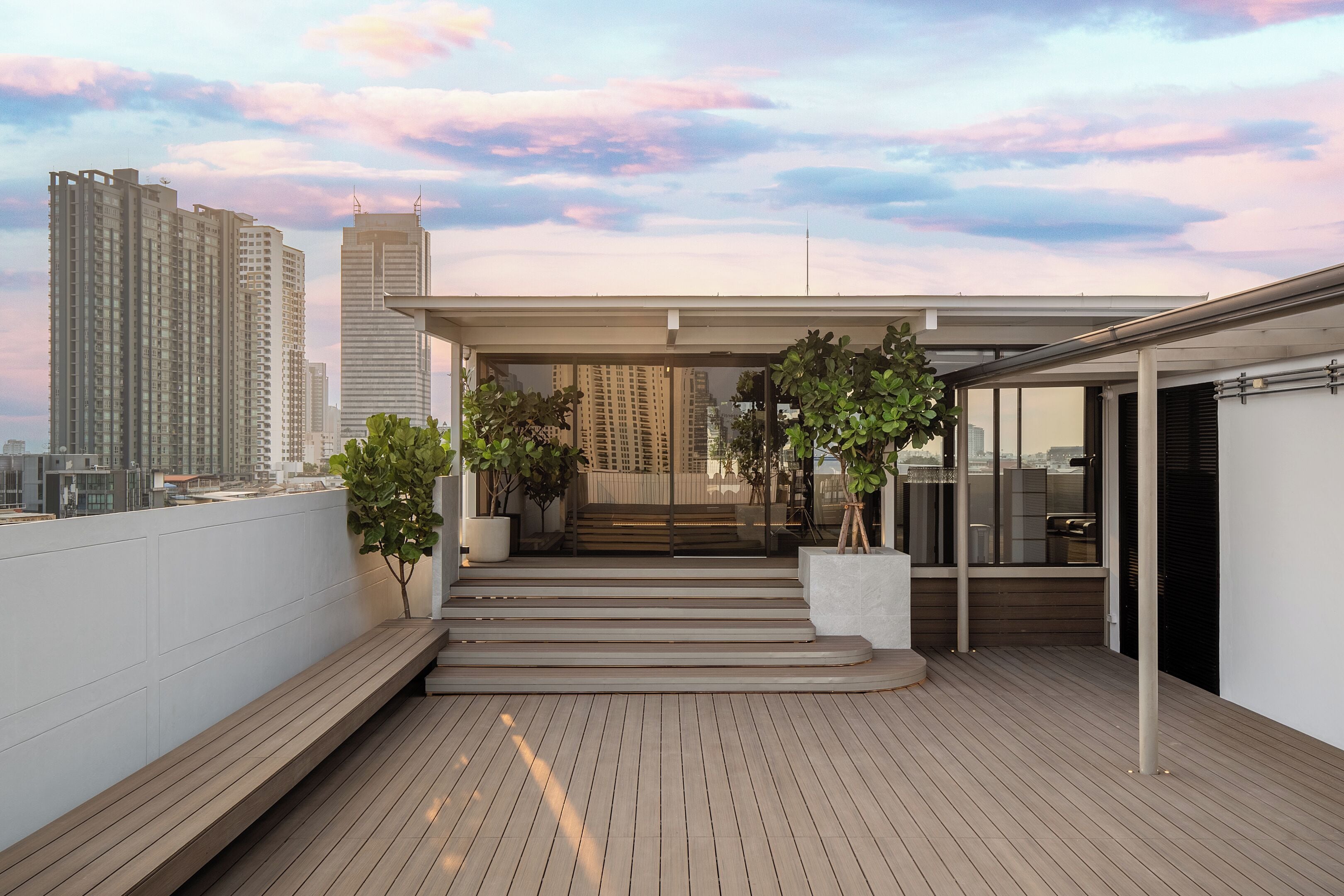 A rooftop terrace with a wooden deck, potted plants, and a view of city buildings.