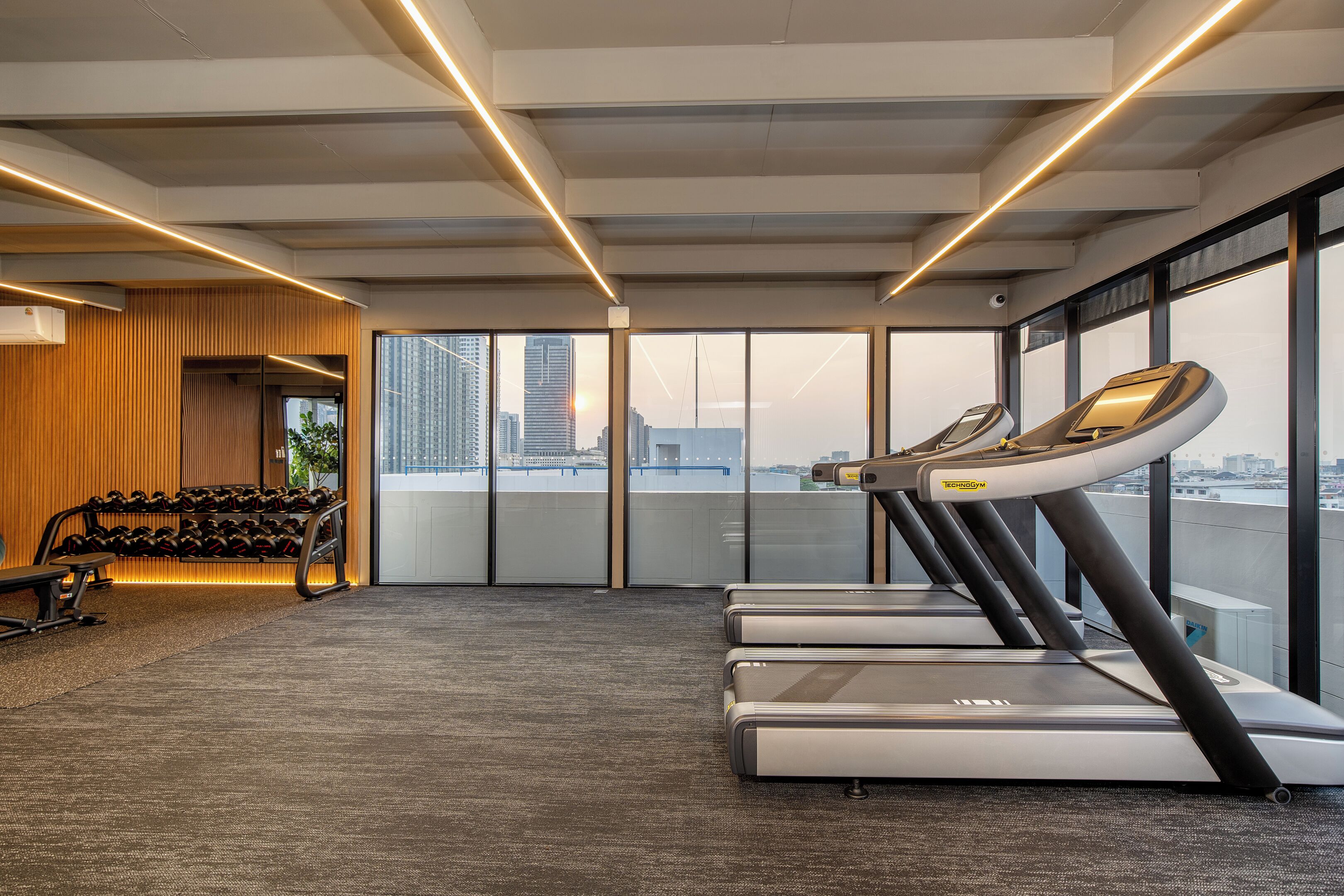 A modern gym with treadmills, dumbbells, and a large window offering a city view.