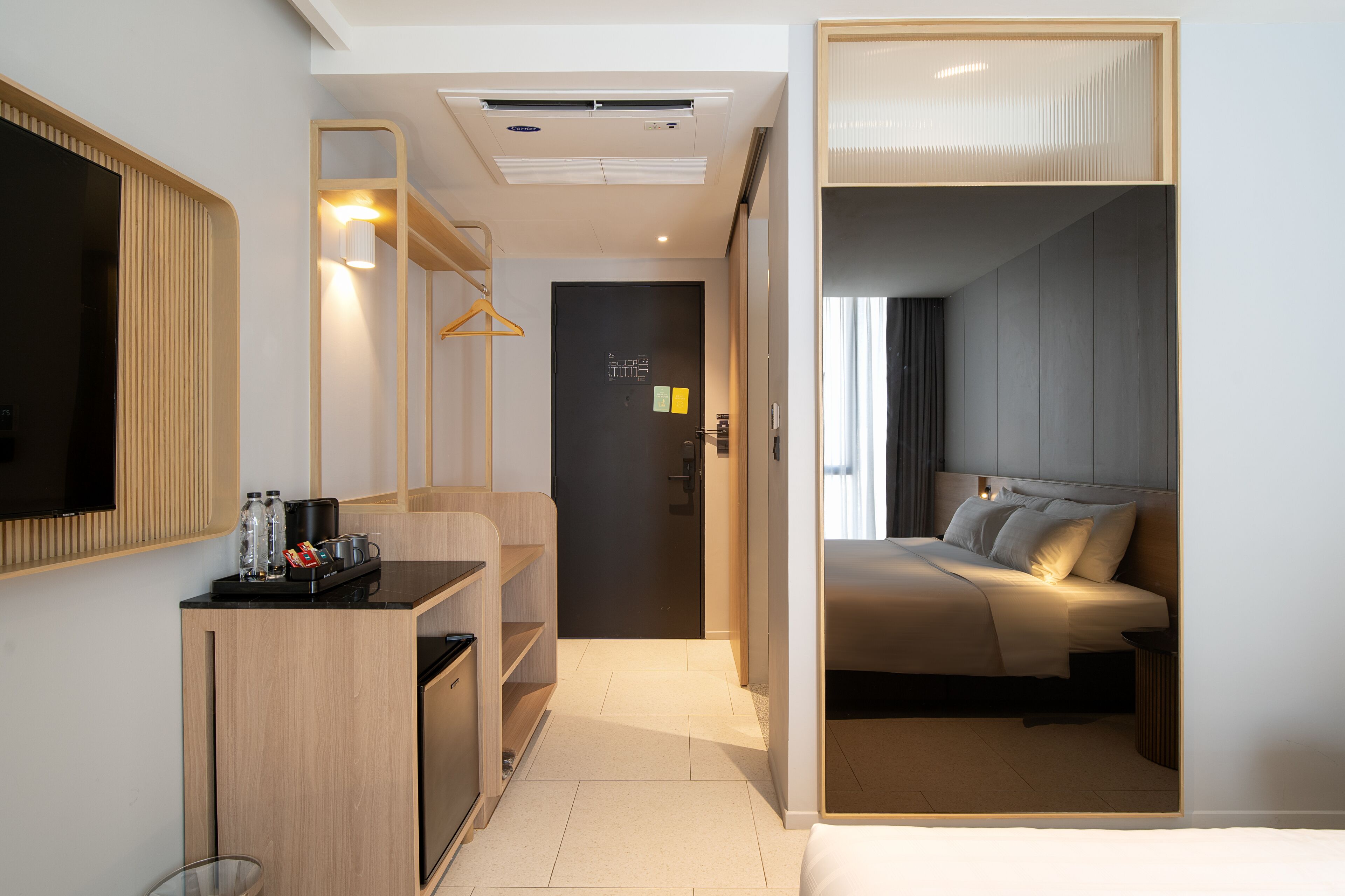 A modern hotel room with a kitchenette, a bed with a headboard, and a large mirror.