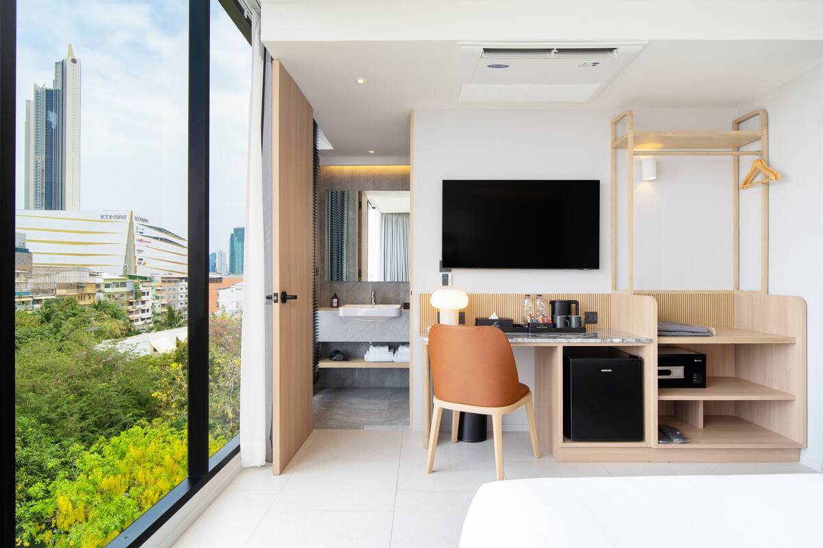 A modern hotel room with a large window, a desk, a chair, a flat-screen TV, a mini-fridge, and a built-in sink.