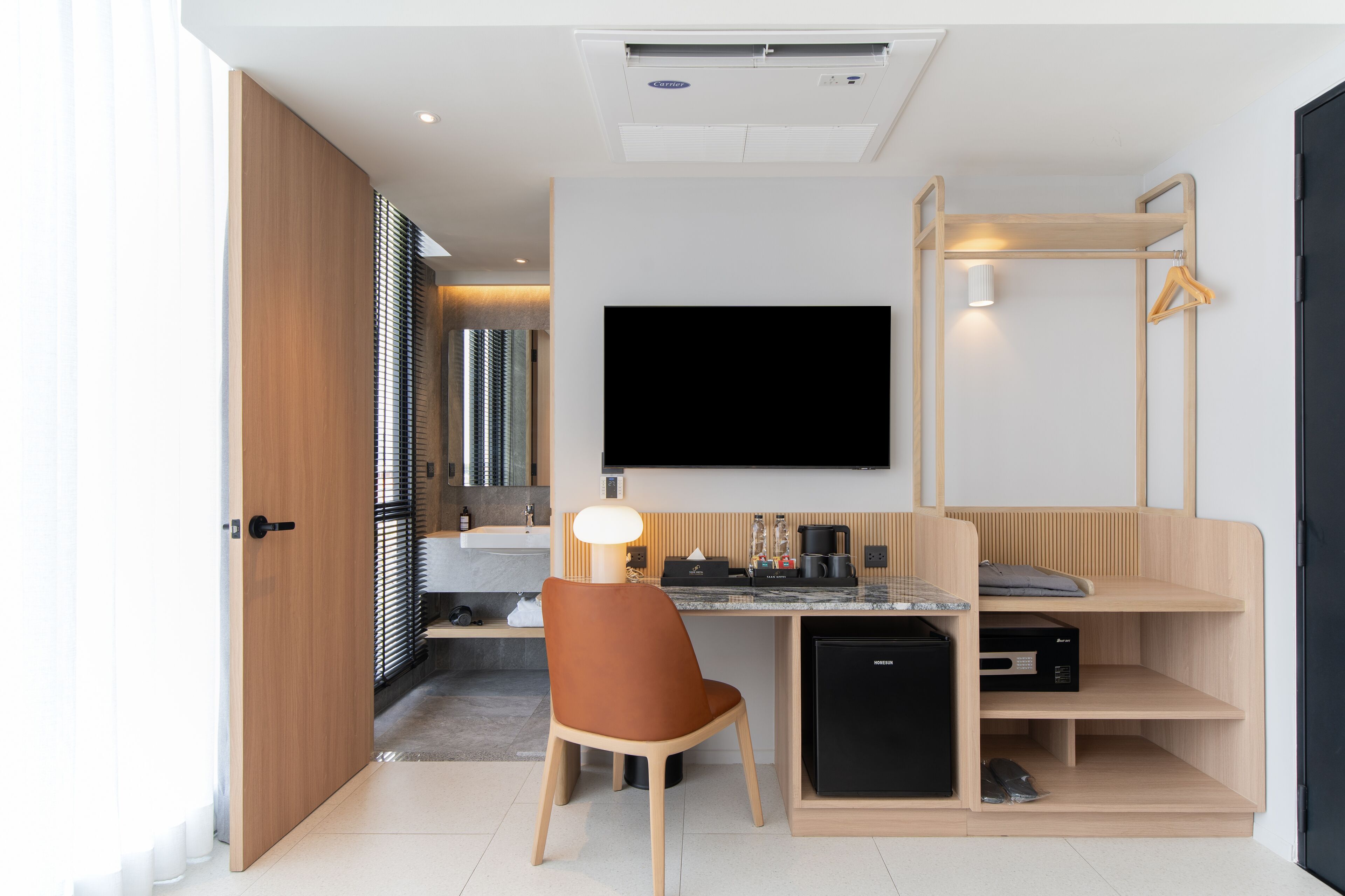 Habitación de hotel compacta con kitchenette, televisor de pantalla plana montado en la pared, una silla de madera y una pequeña zona de escritorio.