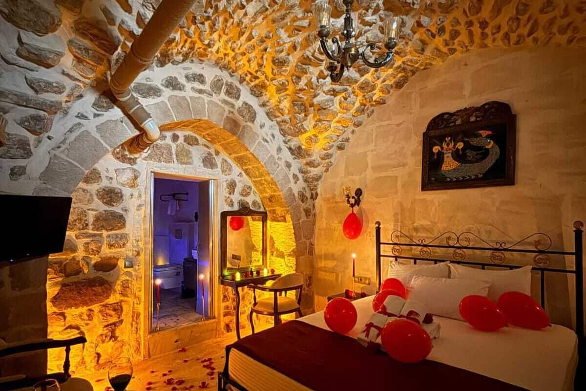 Une pièce avec un plafond en pierre, un lit décoré de ballons rouges, un lustre et un mur en pierre orné d’un tableau encadré.
