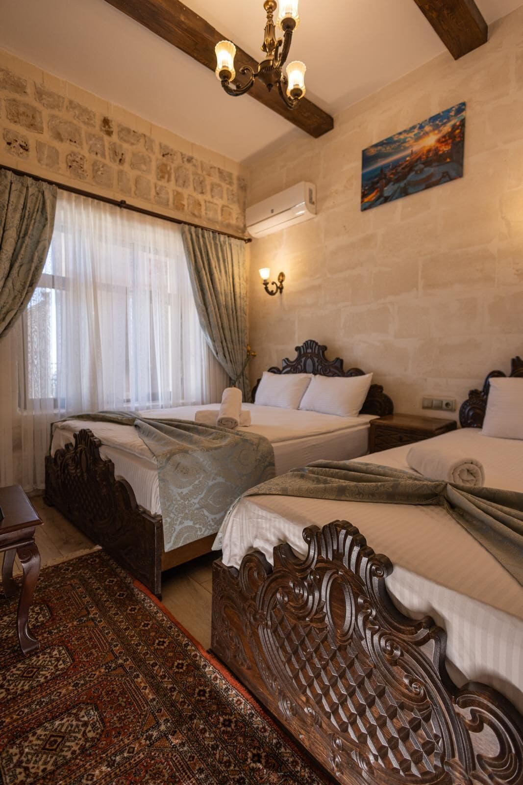 A hotel room with two beds, a patterned rug, a wooden headboard, a chandelier, a painting, and a stone wall.
