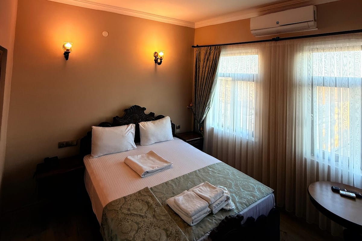 Une chambre d’hôtel avec un grand lit, deux lampes fixées au mur, un appareil de climatisation, une petite table ronde et une fenêtre avec des rideaux.