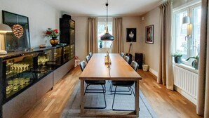 Een eetruimte met een lange houten tafel, stoelen, een kroonluchter in het midden en een keuken met glazen kasten en een fornuis.