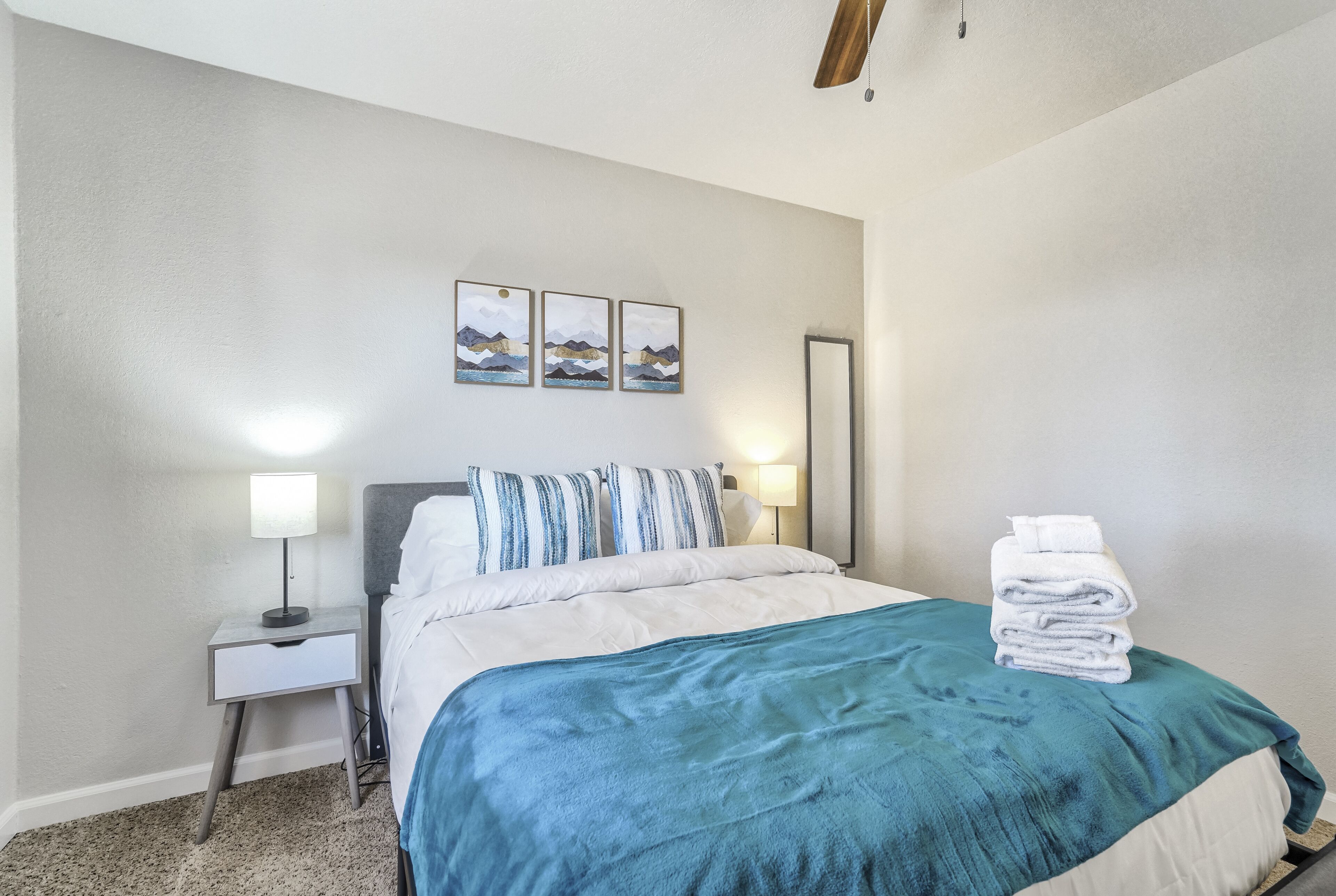 A neatly made bed with a blue blanket, white pillows, and a stack of towels on the side. There are two bedside tables with lamps, and a wall-mounted mirror.