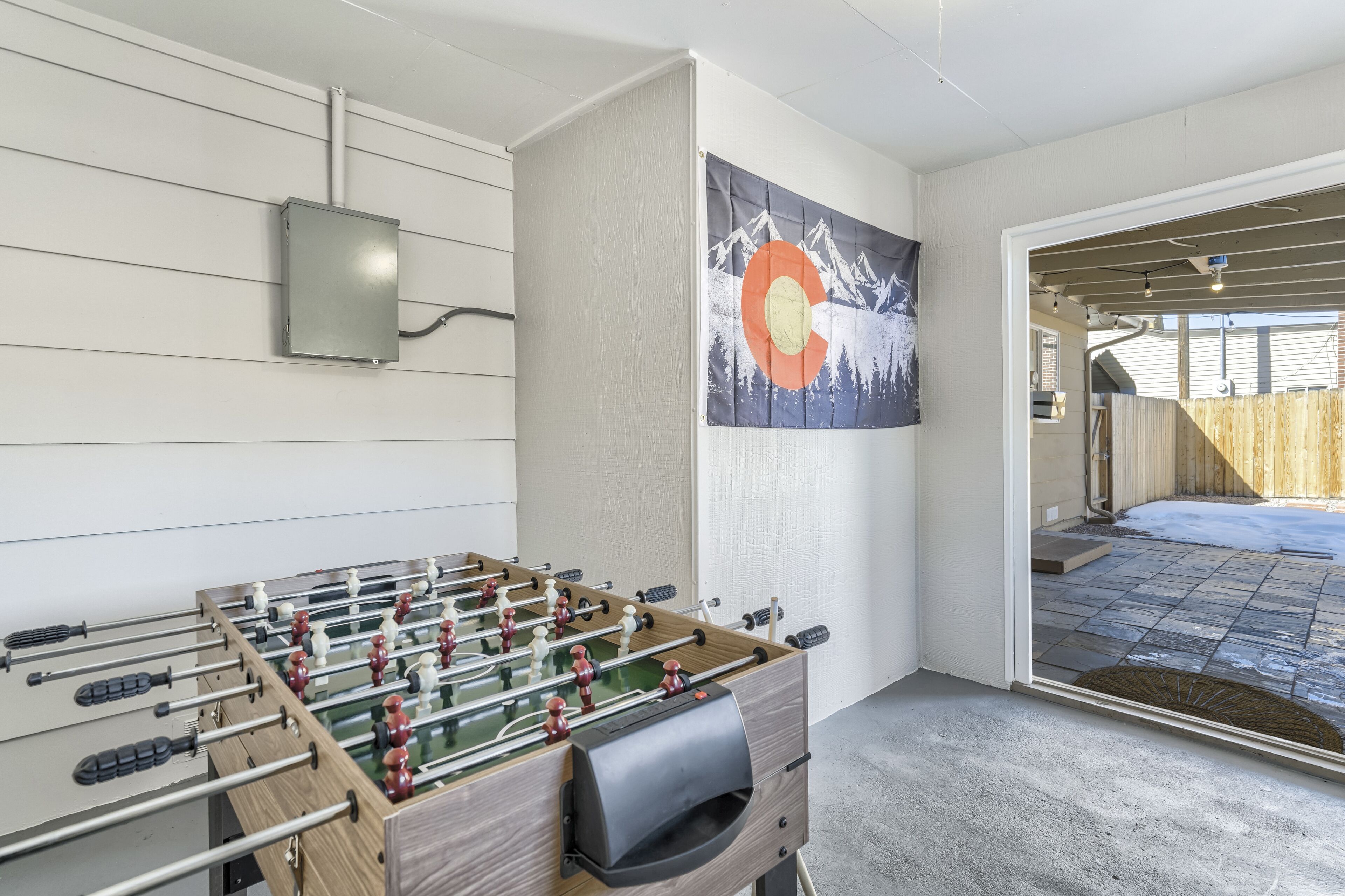 A room with a foosball table, a painting of mountains, and a view of a backyard with a wooden fence.
