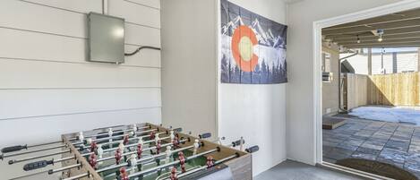 A room with a foosball table, a painting of mountains, and a view of a backyard with a wooden fence.