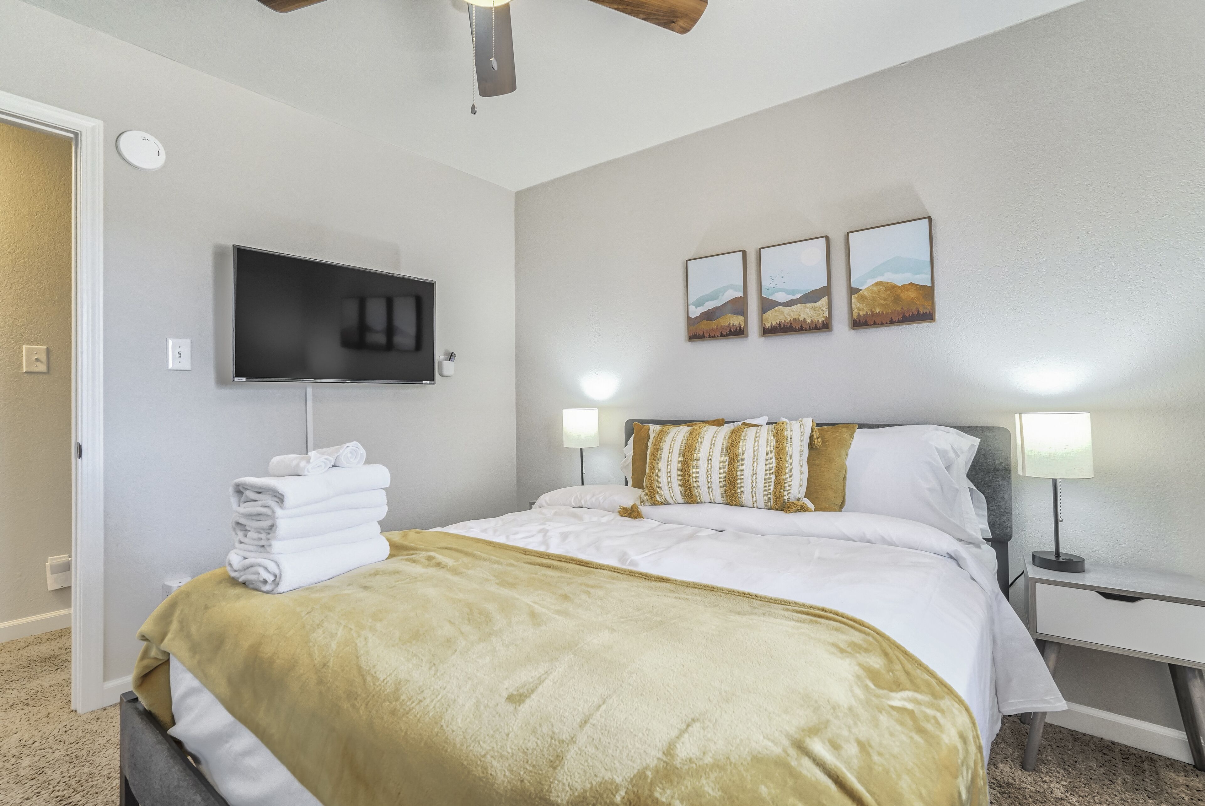 A neatly made bed with a gold throw blanket, a flat-screen TV mounted on the wall, and two bedside lamps.