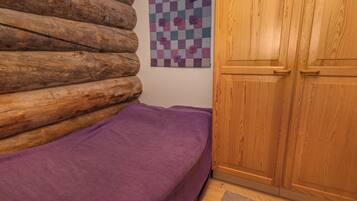 Una habitación con pared de troncos de madera, colcha morada, tapiz a cuadros y armario de madera.