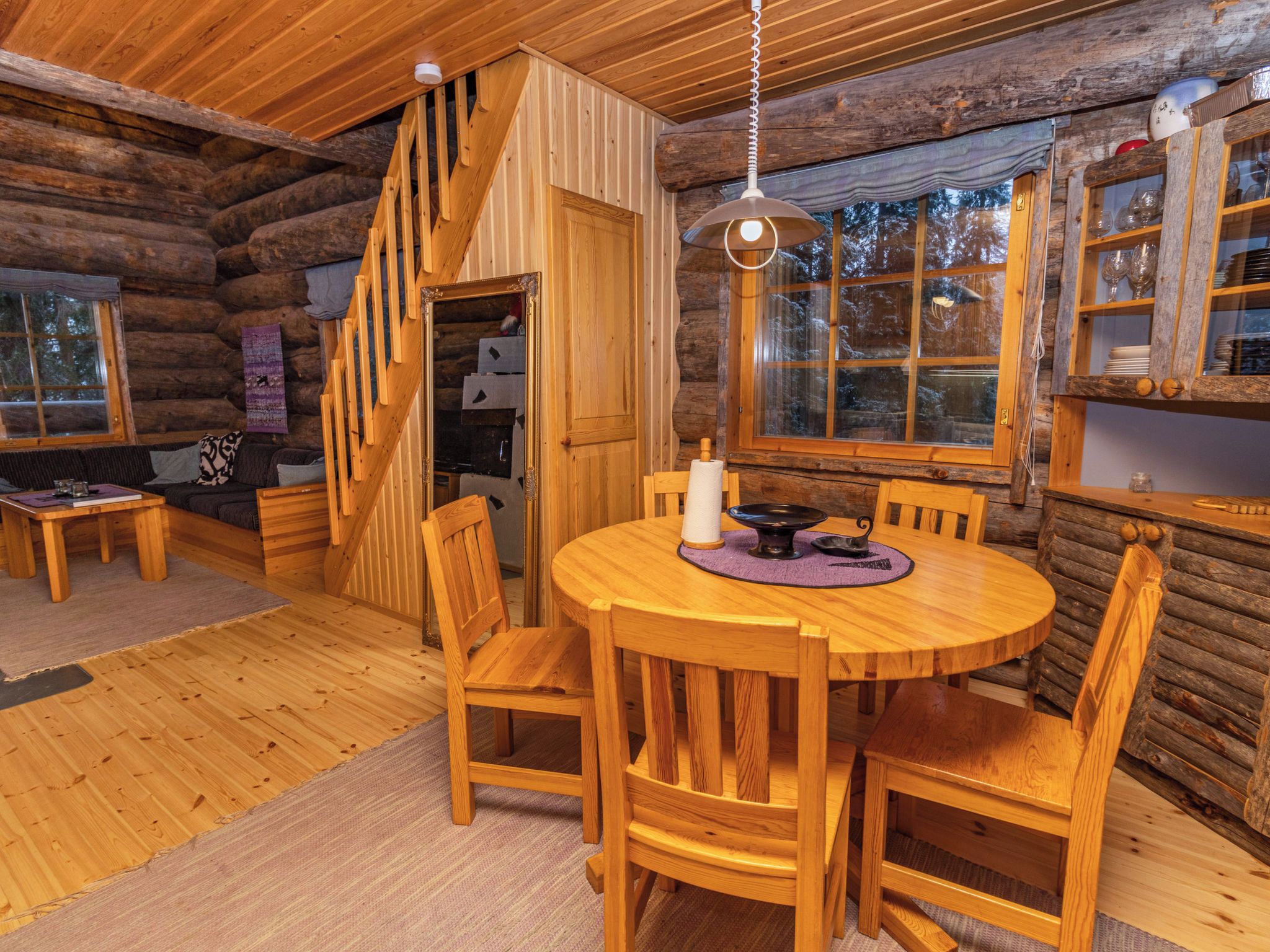 A wooden interior with a round dining table, chairs, a staircase, and a window with a view of trees.